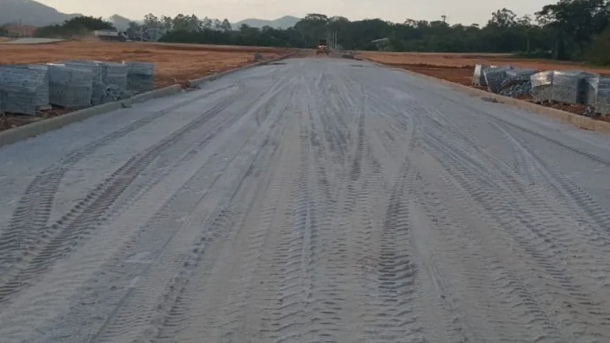 Rua larga do Loteamento Cidade Jardim II, de chão cinza com marcas de pneu, com meio-fio e pilhas de material dos dois lados.