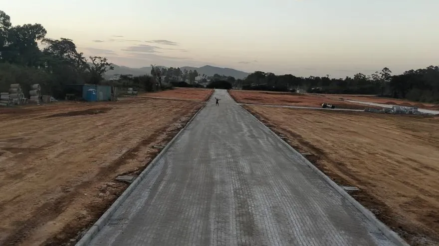 Rua do Loteamento Cidade Jardim II vista em linha reta, com meio-fio dos dois lados, terra exposta nas laterais e morros ao fundo.