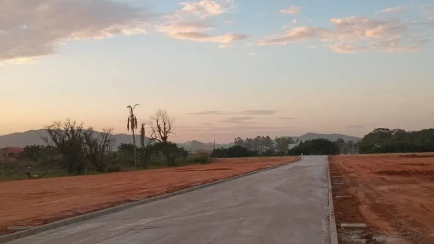Rua do Loteamento Cidade Jardim II, de chão cinza, com meio-fio e terra exposta dos dois lados, árvores e morros ao fundo e céu rosado.