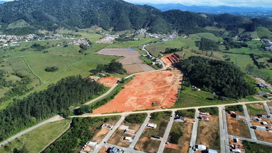 Vista aérea da área do Loteamento Cidade Jardim II: terreno de terra exposta no centro, cercado de verde, com ruas e casas em primeiro plano e morros ao fundo.