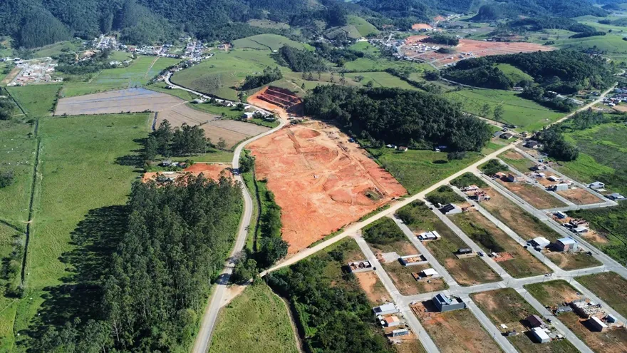 Vista aérea do terreno do Loteamento Cidade Jardim II, de terra exposta, com ruas e construções em primeiro plano e morros ao fundo.